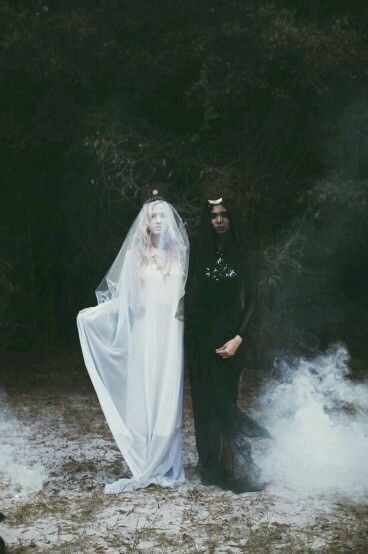 two women in white and black dresses stand in a mysterious forest. the woman in the white dress wears a long veil. the two women appear to be standing in fog or mist. the image has a dark and spooky feeling.