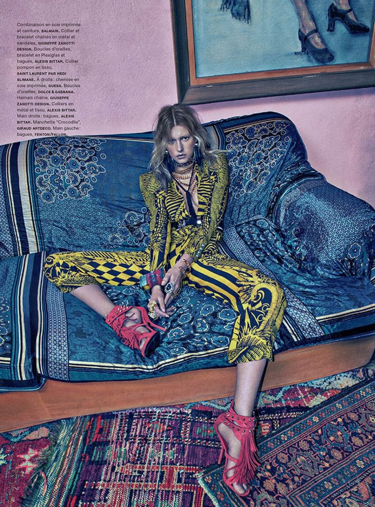 a full shot of a fashion model posing on a richly patterned sofa. the model is wearing a vibrant yellow and black printed jumpsuit with a bold, geometric pattern.  she accessorizes with layered necklaces, bracelets, rings, and anklestrap sandals. the overall style is bohemian chic with a mix of textures and patterns. the setting includes an ornate rug and a piece of artwork in the background, enhancing the luxurious, highfashion feel. the image is styled for a fashion magazine editorial. the text overlay provides details about the designer brands of the clothing and accessories.