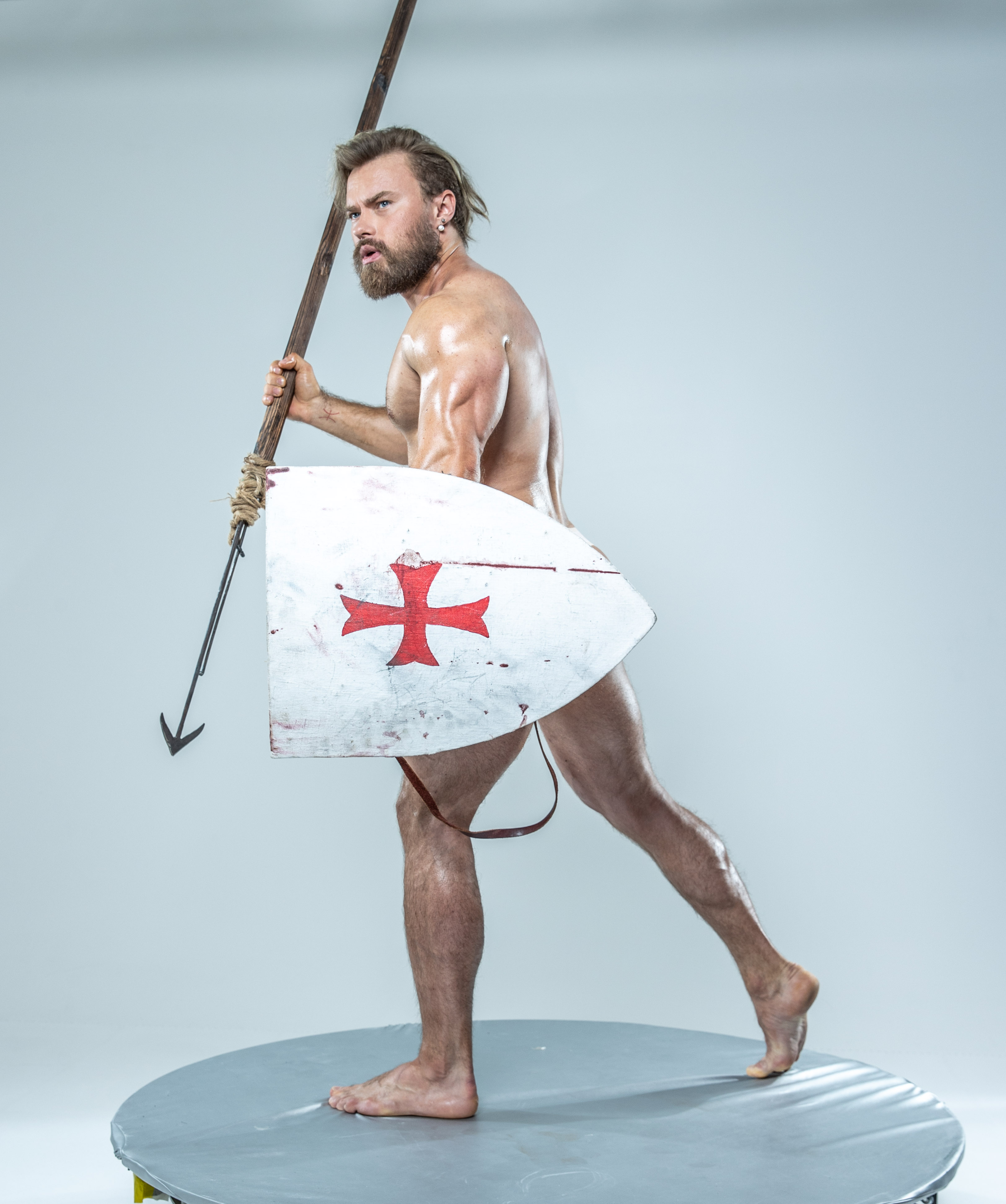 muscular man with a beard and long hair, his body oiled and glistening, stands poised for battle against a seamless white studio background. he wields a spear in his left hand and a weathered shield emblazoned with a red cross in his right, his stance suggesting imminent action. his only attire is a leather strap across his upper thigh, adding to the raw and primal energy of the scene. the studio lighting accentuates his musculature and the texture of his skin, creating a powerful and dramatic image.