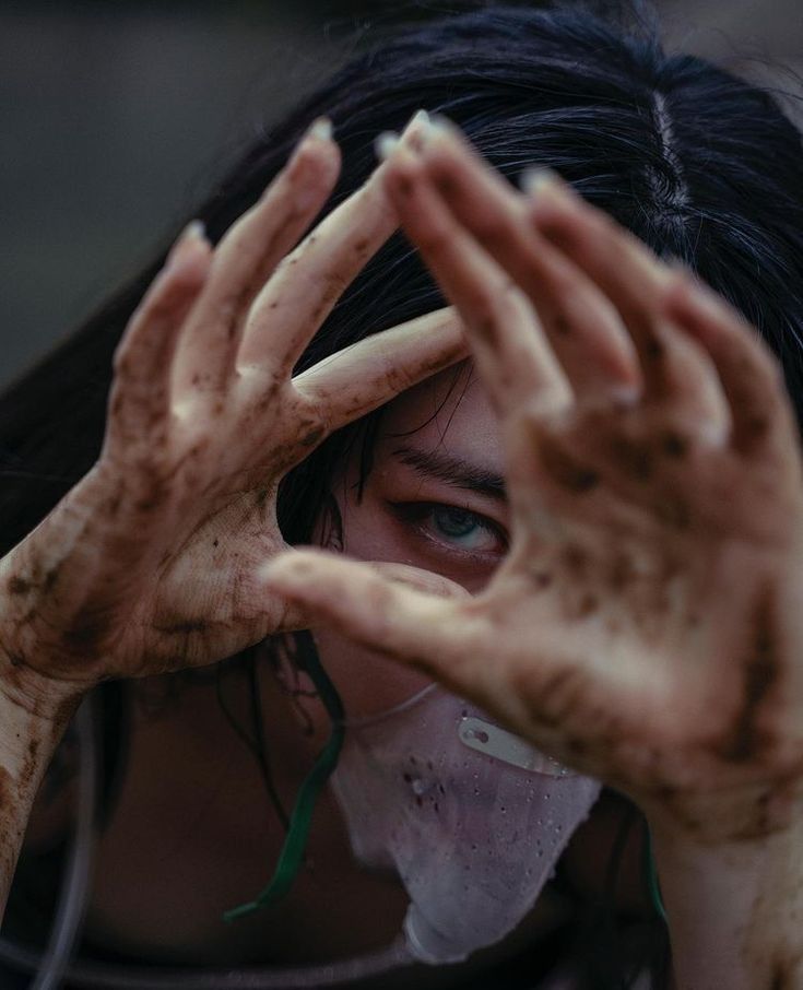 the image is a closeup portrait of a young woman with dark hair, her face partially obscured by her dirty hands held up in front of her. her hands are heavily covered in mud or dirt, emphasizing a sense of grime and struggle. she is wearing a makeshift, damp face mask, further suggesting a postapocalyptic or dystopian setting. her visible eye shows a hint of fear or anxiety. the overall mood is dark, dramatic, and evokes a sense of survival in a harsh environment.  the focus is on the texture of the dirt and the expression in the woman's eye, conveying a story of hardship and possibly environmental collapse.