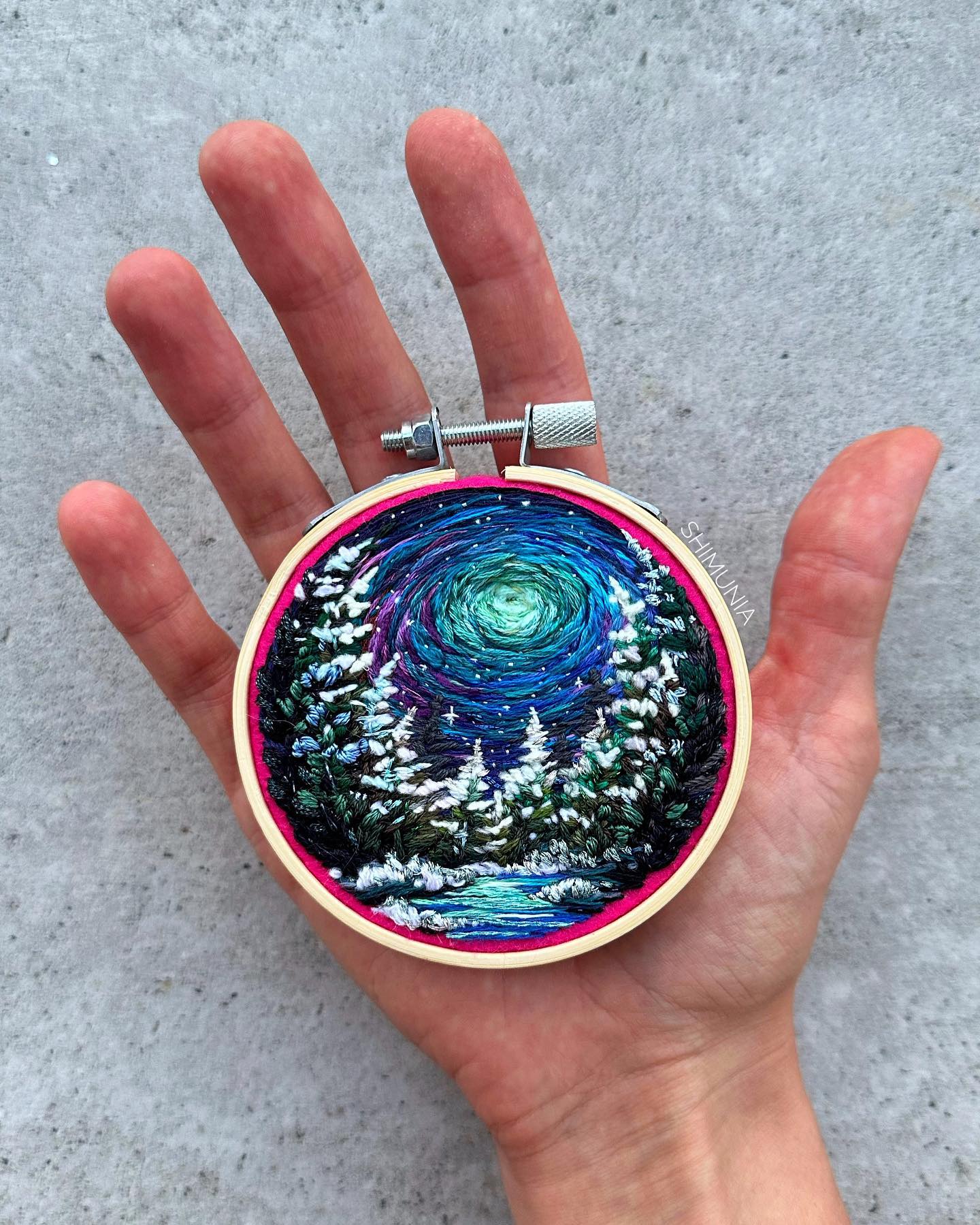 an embroidered artwork of a snowy forest at night is held in an open hand against a gray background. the embroidery is done with various shades of blue, green, and white thread, depicting a serene winter landscape under a vibrant starry sky. the hoop is wooden with a bright pink outer ring and a metal tightening screw.