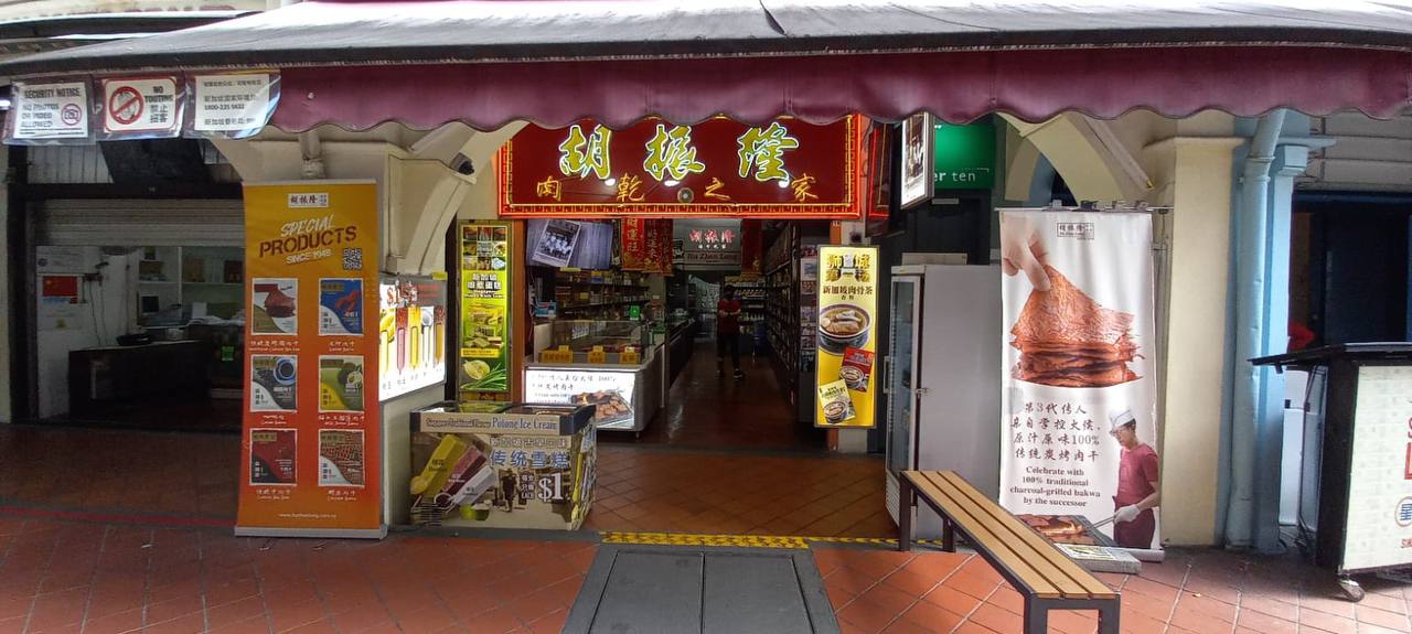 a shot of a storefront in singapore with signage in chinese characters above the door. there are several signs for various food products, including bakwa.  the store appears to be a traditional snack shop selling chinese food, like bakwa.  a wooden bench is in front of the store.
