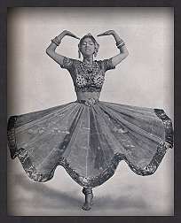 a black and white photograph shows a woman in traditional indian dance attire performing a classical dance. she is wearing a decorative top, a full skirt, and jewelry. her arms are raised and her hands are positioned above her head in a traditional mudra. she is standing on one leg.