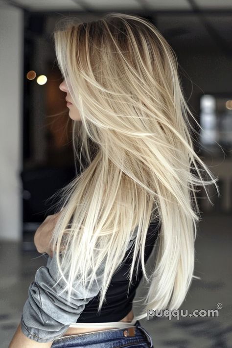 a woman with long, straight, blonde hair styled in a layered cut and ombre color.  she is standing with her back to the camera, looking to her right.  she is wearing a dark gray shirt and blue jeans. the background is blurred.