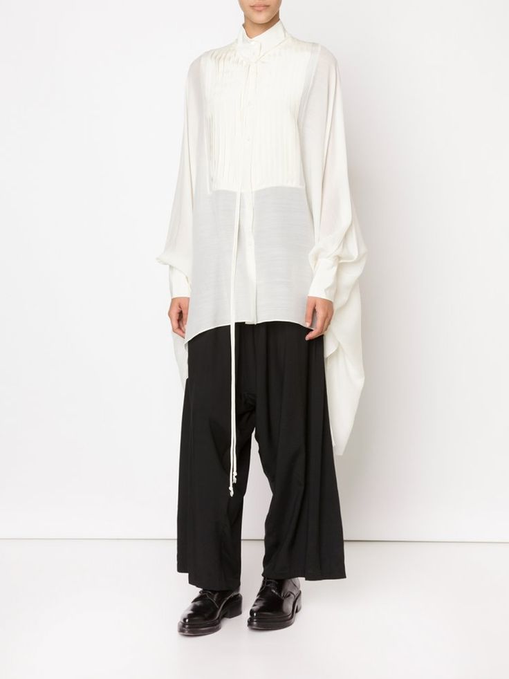 a woman stands in a studio wearing a white, longsleeved blouse and black, wideleg pants. she is also wearing black dress shoes. the blouse has a pleated front and high neck. the pants are highwaisted and have a drawstring. the background is white. the image is a full shot.
