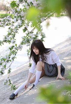 an asian woman wearing a student uniform sits outdoors with flowers and tree branches around her. she is wearing a white shirt, gray skirt, white socks, and black loafer shoes. she appears to be adjusting her shoe.