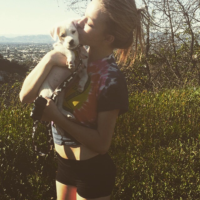 the image shows a young woman with blonde dreadlocks pulled back into a ponytail, wearing a tiedye shirt and black athletic shorts. she's outdoors on a hillside overlooking a city (possibly los angeles), bathed in sunlight.  she's holding a small, white puppy and is kissing it affectionately on the head.  the background includes green bushes and a view of a city in the distance.  the overall mood is happy and affectionate.