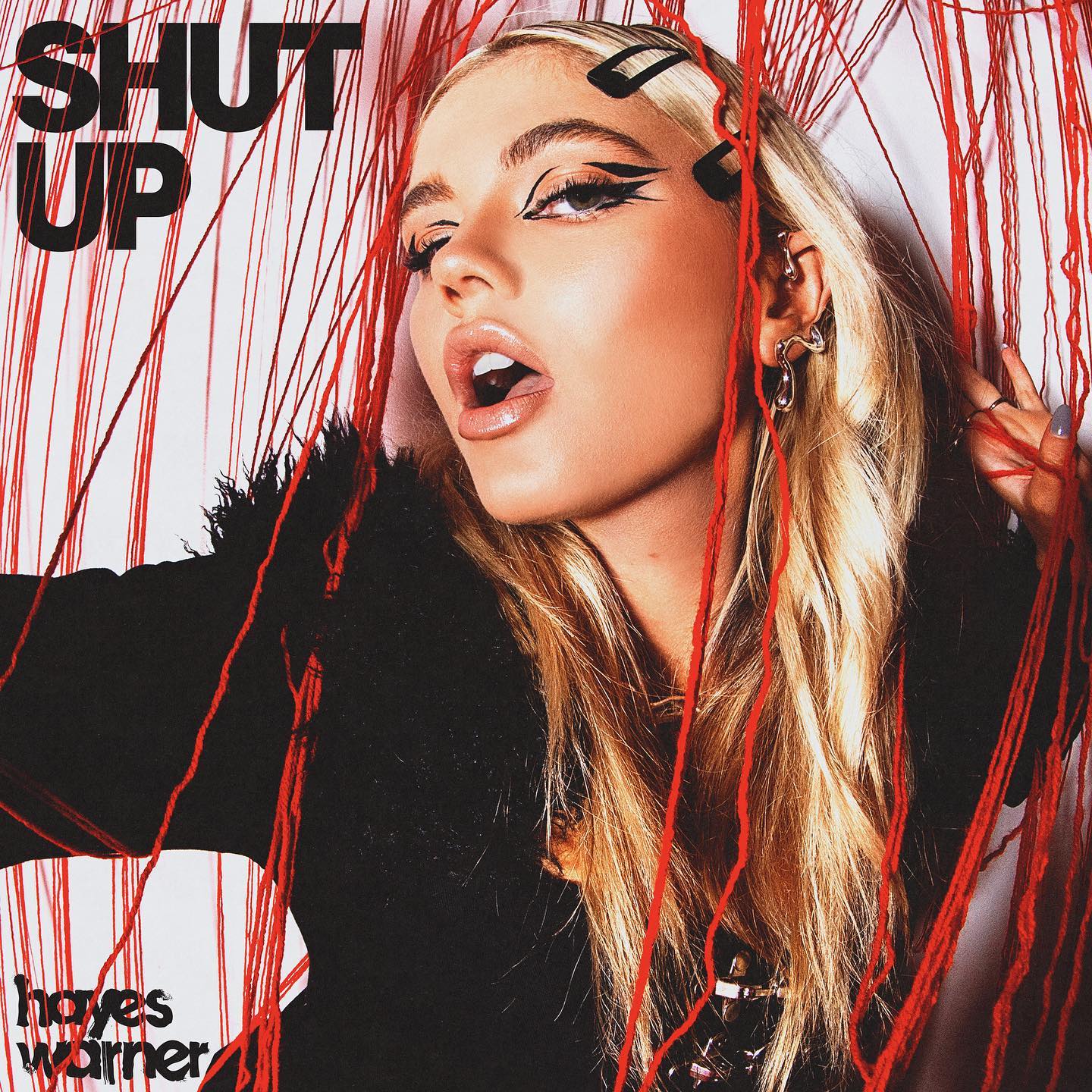 an album cover features a woman with blonde hair and black hair clips, wearing winged eyeliner, and various pieces of jewelry. she has her mouth open, and red strings surround her. the artist's name, hayes warner, is in the bottom left corner and the album title, "shut up", is in the top left corner.