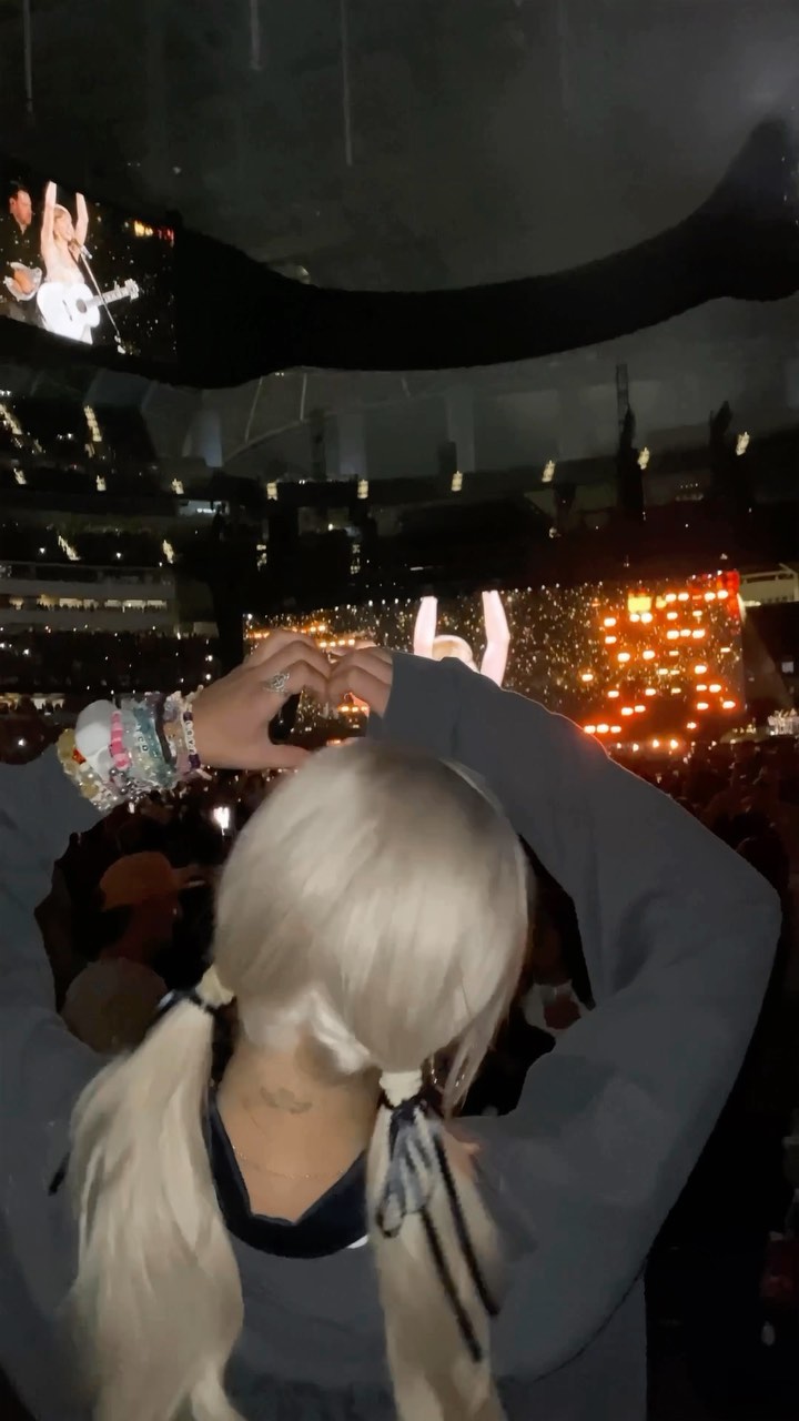 a concert photo shows a person with blonde hair in pigtails making a heart hand gesture in front of a litup stage with a crowd. the person is wearing bracelets. a screen showing the performers is also visible.