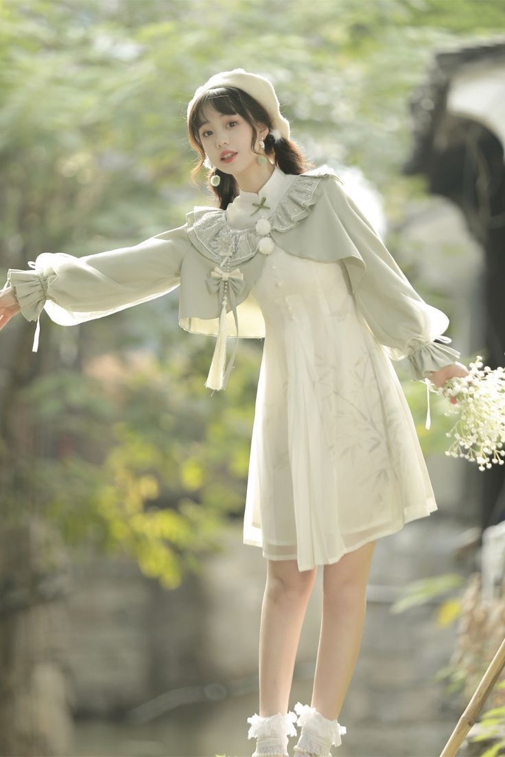 a young woman stands on a stone bridge, posing for the camera. she is wearing a flowing, white lolita fashion dress with a green capelet. her outfit is accented with delicate embroidery and ruffles. she holds a bouquet of small white flowers in her left hand. the background features lush greenery and a glimpse of water, suggesting a serene and natural setting. the lighting is soft and natural, casting a warm glow on the scene.