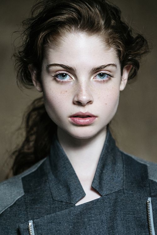 the image is a portrait of a young woman with pale skin and freckles. she has long brown hair and blue eyes. she is wearing a gray jacket with a high collar. the background is a neutral brown color. the lighting is soft and natural, creating a warm and inviting atmosphere. the woman's direct gaze and neutral expression give her a sense of confidence and composure.