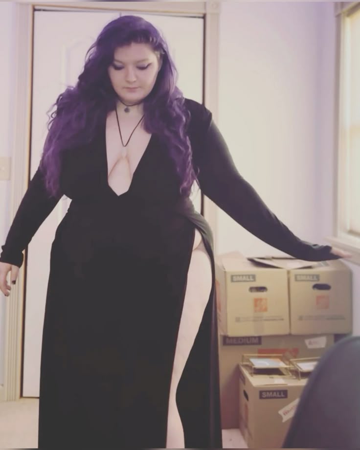 a plussize woman with purple hair is wearing a long black dress with a plunging neckline and a high slit on one leg. she is standing in a room with moving boxes.