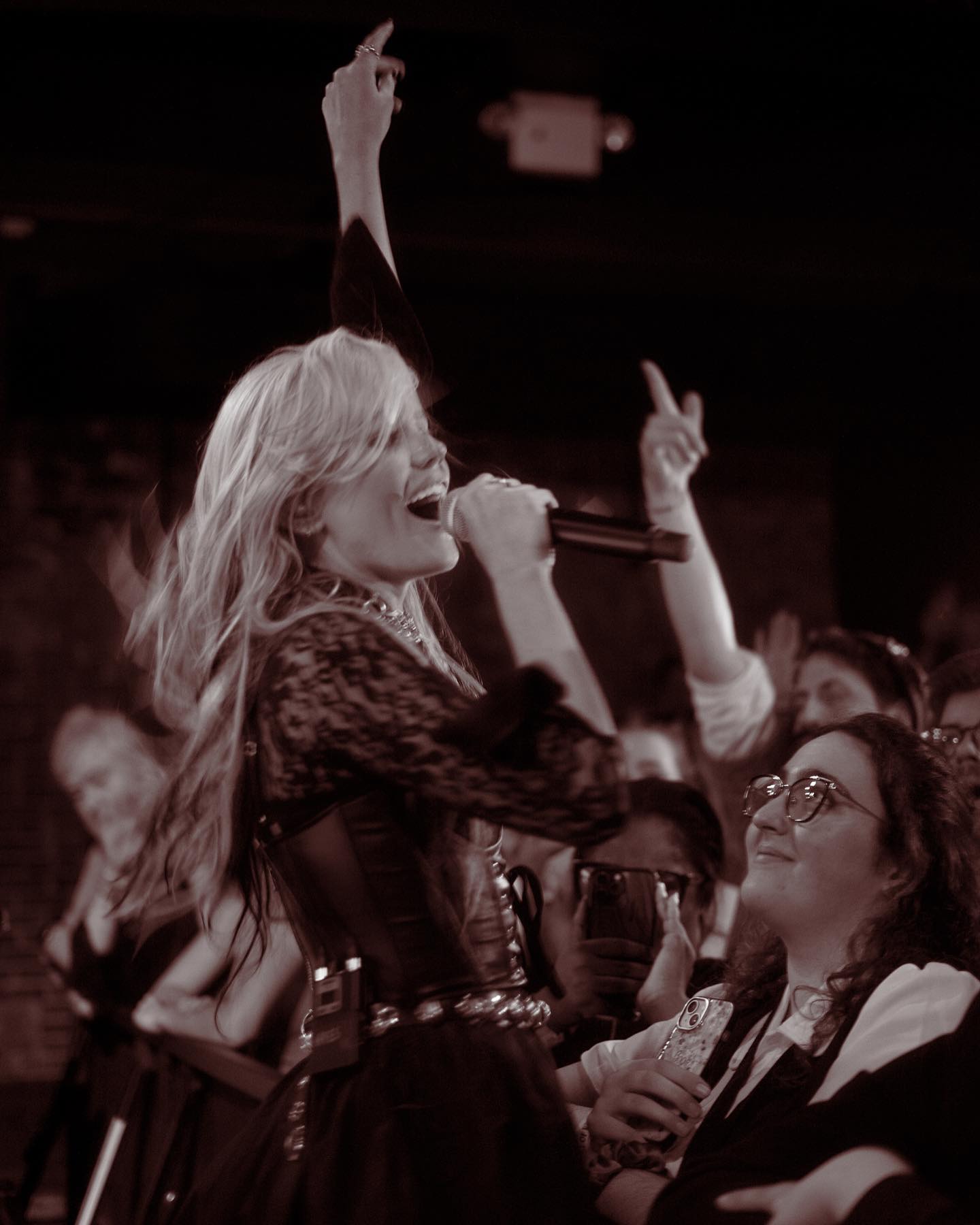 a sepia toned photo shows a female singer on stage at a live concert. she has blonde hair and is wearing a dark, possibly leather, corsetlike garment with a lacelike top and dark skirt. she is holding a microphone and singing with her mouth open. her right arm is raised high, pointing towards the audience. the crowd is visible in the background, some with their hands raised, and others looking up at the stage with expressions of enjoyment and excitement.