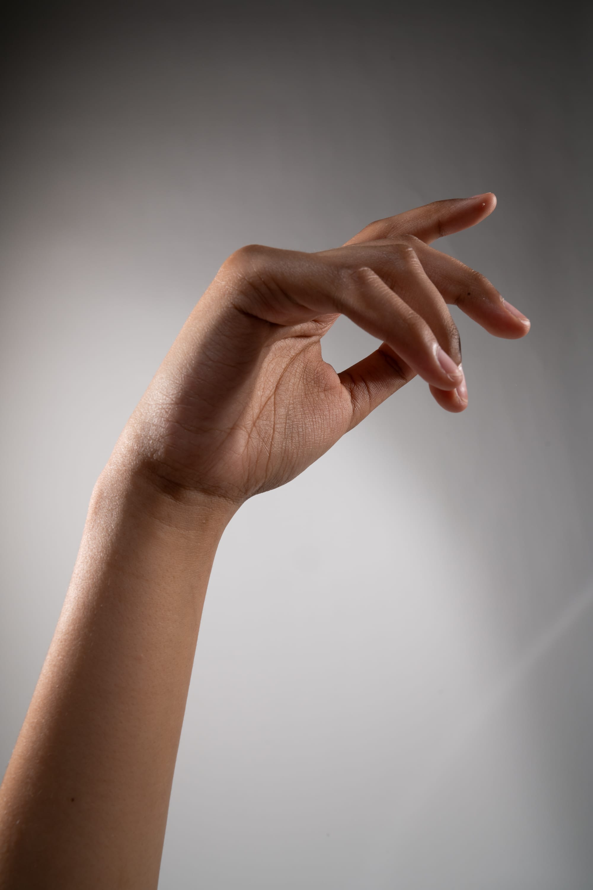 a hand is reaching out with a pinching gesture, as if to grab something small. the skin is medium brown and the fingers are slender. the background is a plain, offwhite color, and the lighting is soft, highlighting the hand and arm.