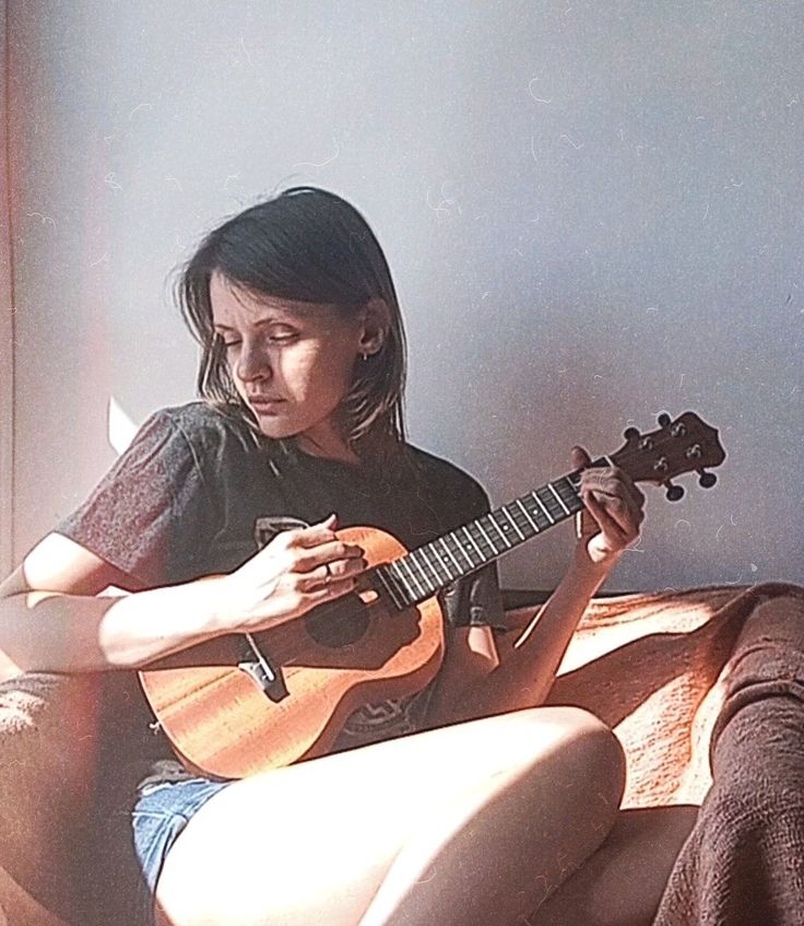 a young woman with brown hair, wearing a black tshirt and denim shorts, sits on a couch playing a ukulele. the woman is smiling softly and seems relaxed. sunlight streams in from the window to her left.