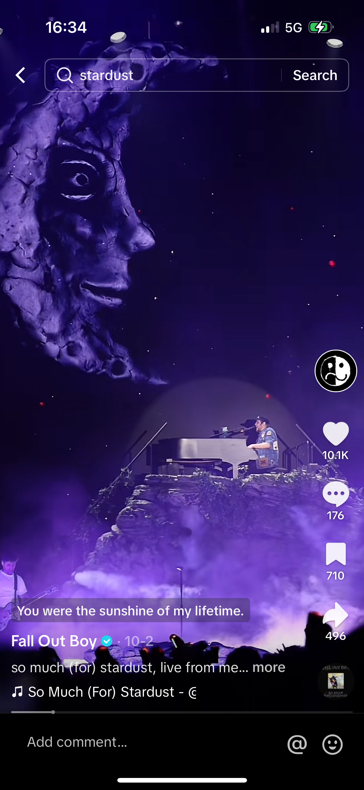 a still image of a fall out boy concert captured from a mobile phone. the band is performing on stage with the lead singer at the piano. the image is dark with purple and blue lighting. the phone screen shows the song title, "so much (for) stardust" by fall out boy. the band is playing to a large crowd, but only silhouettes of the audience are visible. the phone screen also displays various social media features with comment count, likes, shares, and replies.