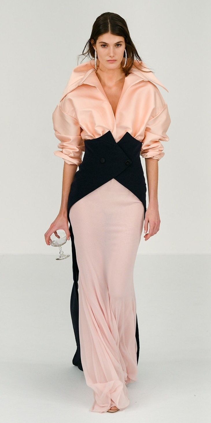a woman in a pink and black dress is posing on a white runway. she has on a pink blouse with a black skirt that is open in the back. she is also wearing black heels and diamond earrings. she has her hair pulled back and is looking straight ahead. the background is white.