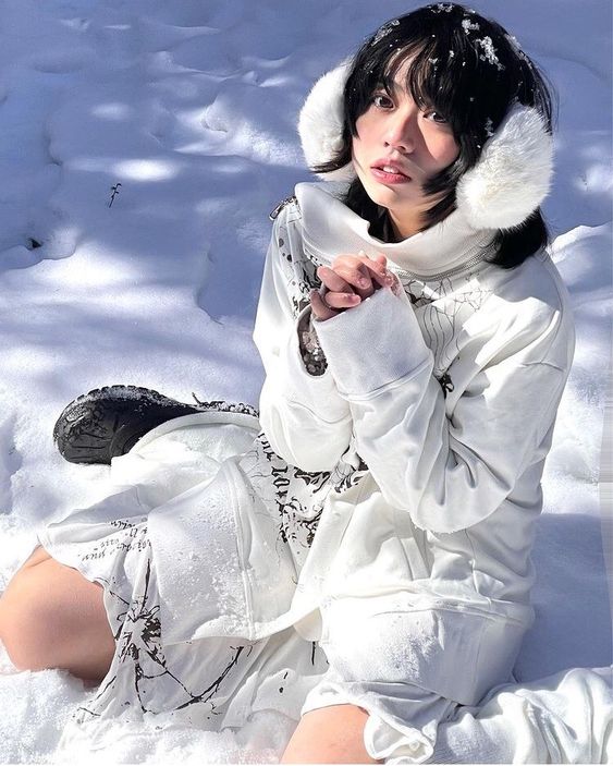 a young woman in a white sweater and earmuffs sits in the snow and looks up at the camera.  she is wearing a white jacket over a white shirt and the entire outfit is white.  she has dark hair and looks at the camera with a serious expression.  there is snow all around her.