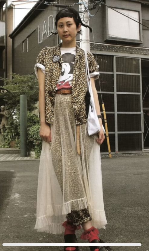 a woman stands outside on the street. she is wearing a tshirt with a mickey mouse graphic on it, and a leopard print buttonup shirt. she is also wearing a leopard print maxi skirt with a layer of tulle on top, and red socks and black sandals. she has short dark hair styled in braids. she is standing in front of a building.