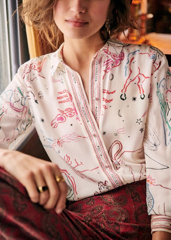closeup view of a woman wearing a sezane longsleeved blouse. the blouse is offwhite or creamcolored and features a whimsical print of constellations, zodiac signs (possibly), animals (including a horse and a scorpion), and other celestial elements. the print is done in muted colors of pink, red, and blue. she is also wearing burgundy paisley print pants. the overall style is bohemian or vintageinspired. the focus is on the detail and texture of the blouse and the woman's hands. the image is welllit, showcasing the intricate design of the shirt.