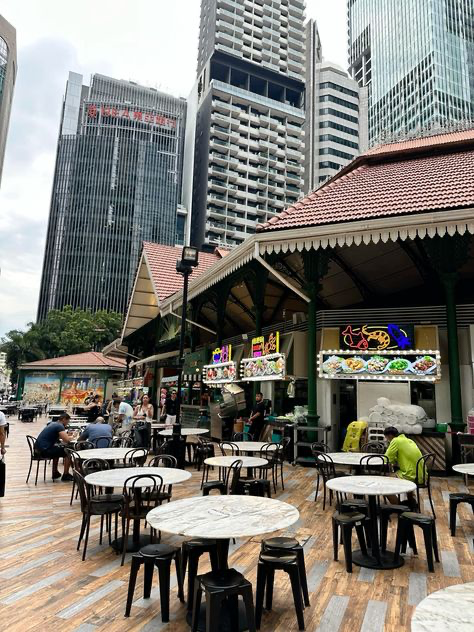 the image shows lau pa sat (telok ayer market) in singapore, a historic victorianera market that now houses a large hawker centre. the photo is taken from the outdoor seating area, showing numerous marbletopped tables and metal chairs arranged across a wooden deck.  in the background, modern skyscrapers rise above the market, showcasing the contrast between old and new architecture in singapore's city skyline.  the sky is overcast. several people are visible, either seated at the tables or walking around the market. the food stalls with signage for various asian dishes are partly visible behind the seating area.  the overall atmosphere is that of a busy, yet relaxed, outdoor dining area in a bustling urban setting.