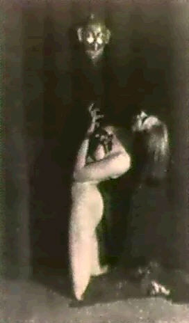 a black and white photograph depicting a dramatic scene. a nude woman kneels before a shadowy figure, possibly a man wearing a mask, that resembles a devil or demonic entity. the lighting is dramatic, with much of the scene shrouded in darkness, adding to the mysterious and suspenseful atmosphere. the overall style is gothic and reminiscent of horror or ritualistic imagery. the image has a vintage or antique feel.
