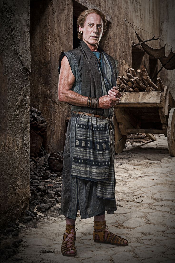 a promotional image of an actor portraying a character in a historical drama, possibly set in ancient times. the man, appearing older with long gray hair, wears a sleeveless tunic and a patterned apronlike garment over it, along with sandals. he stands in a street of an ancientlooking city, with a cart and a pile of wood in the background. the overall tone of the image suggests an historical or mythological setting.  the actor's serious facial expression adds to the dramatic quality of the portrait. the image style and quality suggest it may be a still from a television show or movie.