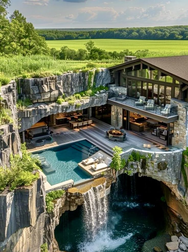 an aerial view of a modern house built into the side of a cliff with a waterfall cascading into a pool of water below. the house features a deck with outdoor seating and a pool with lounge chairs. the surrounding landscape includes green fields and trees.