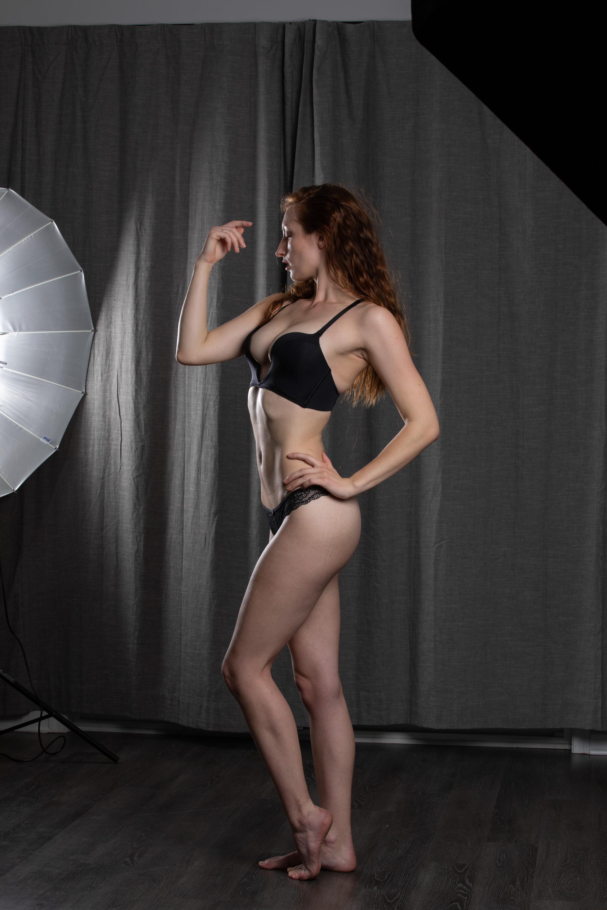 a young, slender woman with long reddish hair poses in a black bra and underwear set against a dark grey backdrop in a photography studio. a large softbox illuminates her from the left, creating soft shadows and highlighting her toned physique. she stands in profile, her left hand lightly touching her forehead and her right resting on her hip, showcasing a confident posture. the overall aesthetic is minimalist and focused on highlighting the model's form.