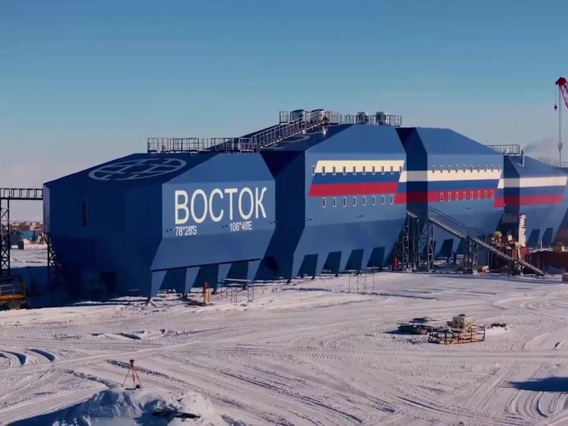 a blue building with "восток" written on it stands in the middle of a snowy, frozen landscape. the building has the russian flag painted on the side of it. it is likely the vostok station in antarctica, a research station run by russia.