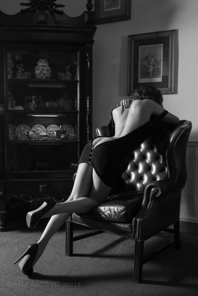 a black and white photo of a woman sitting on a chair in a dark, mysterious bedroom. she is wearing a black dress and high heels, and her pose is