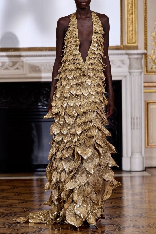 a darkskinned model stands on a stage, wearing a sleeveless, gold, metallic, sequined gown. the gown is embellished with scalelike sequins from the halter neck all the way down to the floor, and extends into a slight train. the gown has a deep vneck. the model is in front of a fireplace and decorative architectural molding.