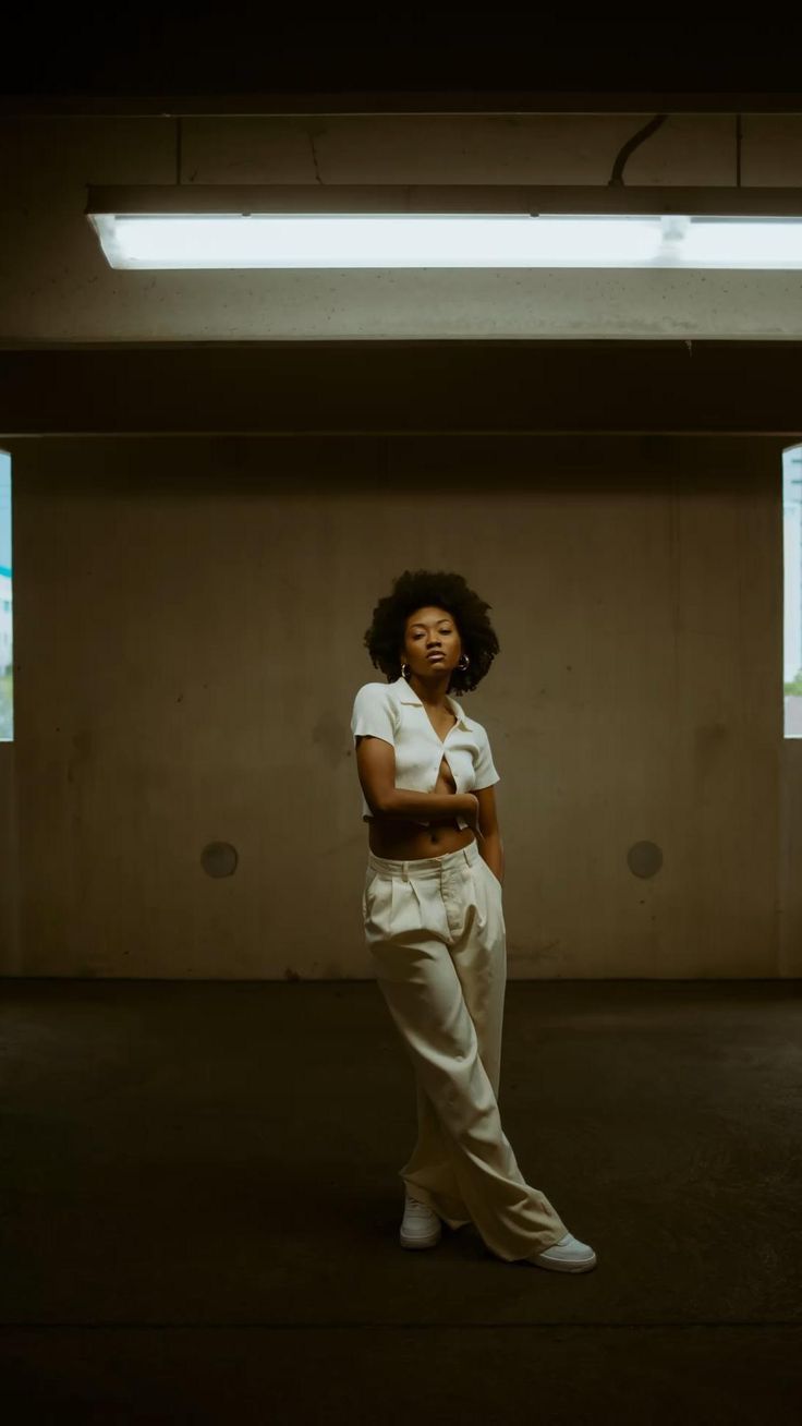 a young woman with dark hair is standing with her legs crossed in a white shirt and matching trousers, in a dimly lit indoor space with a single fluorescent light. she is looking towards the camera with a serious expression. the concrete wall and floor are plain and simple, adding to the minimalist aesthetic of the image.