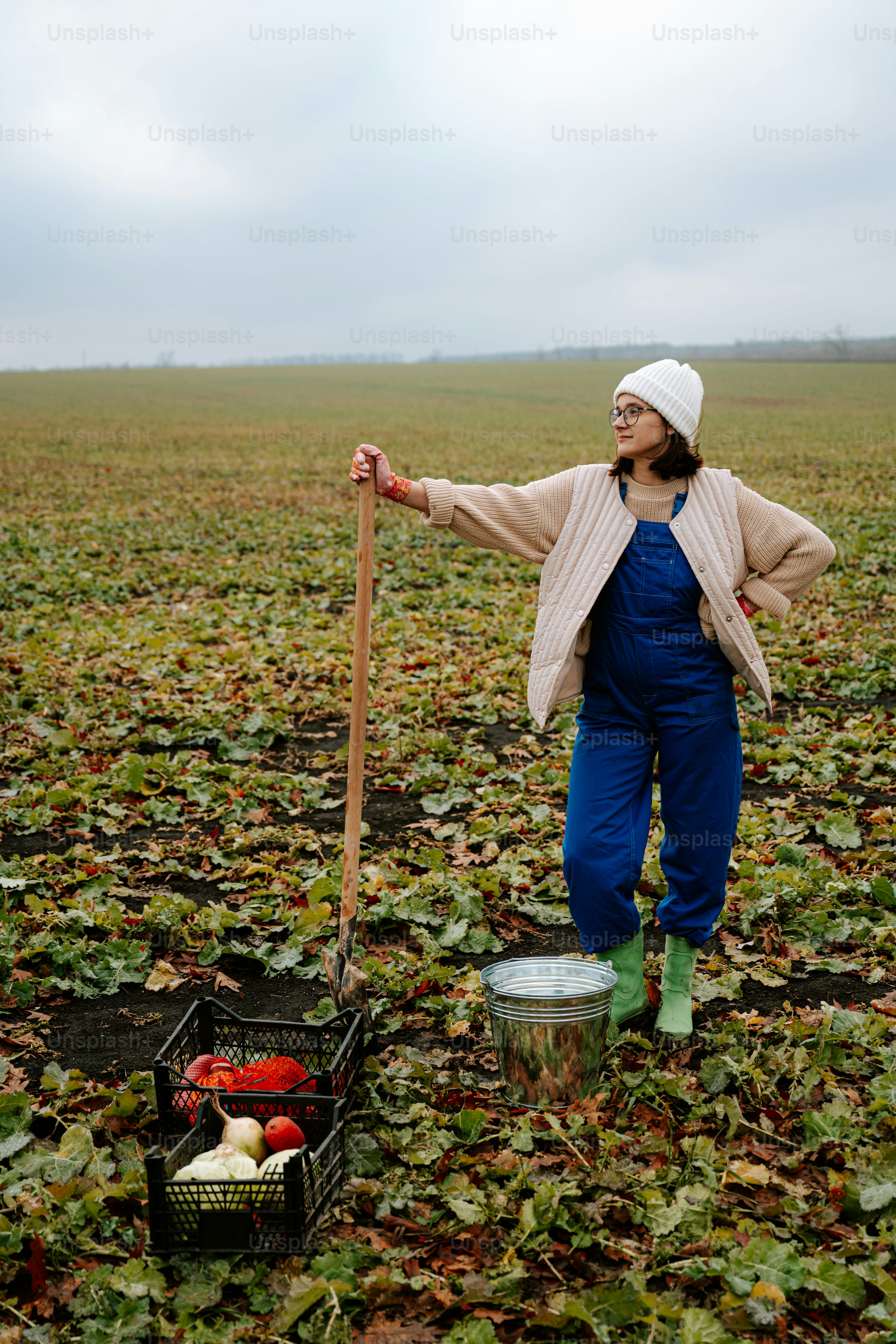 a woman in overalls and a white hat stands in a field of harvested crops, leaning on a shovel.  she has a crate of pumpkins and gourds at her feet and a bucket nearby. the field is covered with fallen leaves, indicating it's autumn. the sky is overcast and the overall mood is peaceful and rustic.