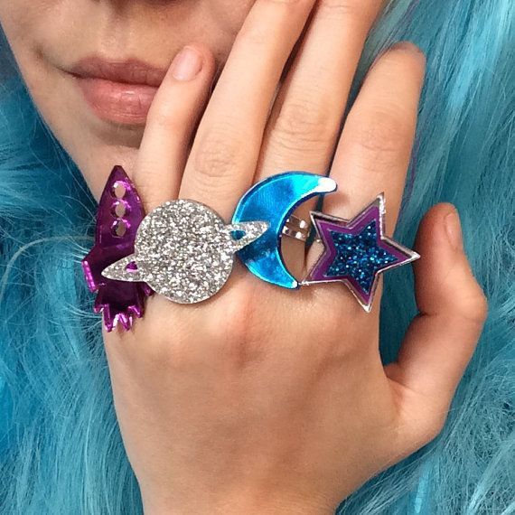 a closeup shot of a hand wearing multiple rings featuring spacethemed designs such as a rocket, planet, moon, and star, all adorned with glitter. the person has blue hair.