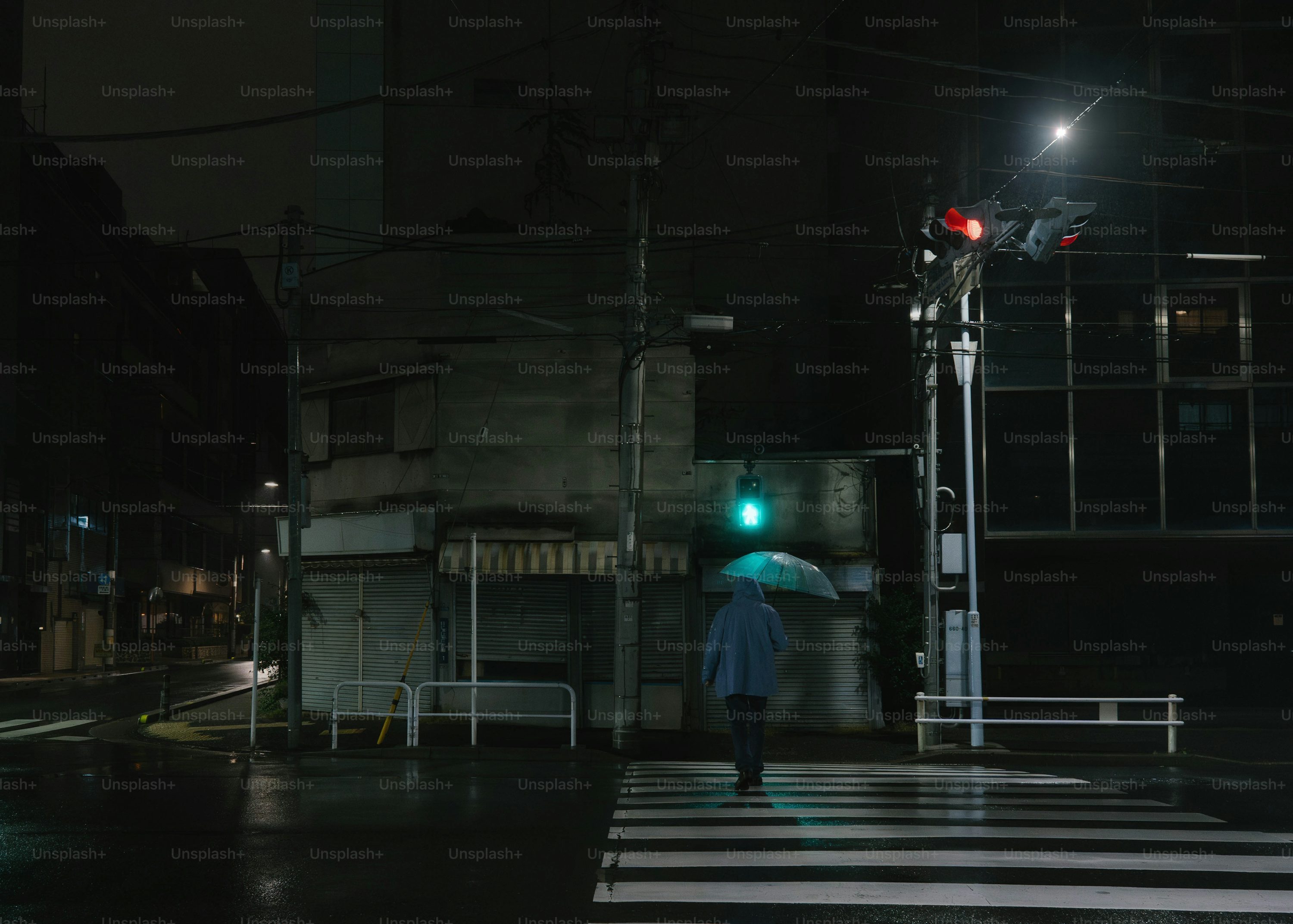 a person with a light blue umbrella is walking across a zebra crossing on a rainy night street. the streetlights are on, illuminating the wet asphalt. traffic lights are visible, with one showing green and the other red. buildings line the street in the background.