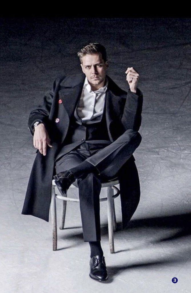 a man in a black suit and coat is sitting on a chair. he is looking at the camera with a serious expression on his face. he is wearing black shoes. the man's legs are crossed and he is wearing a white shirt under his suit jacket. the background is a dark gray color.
