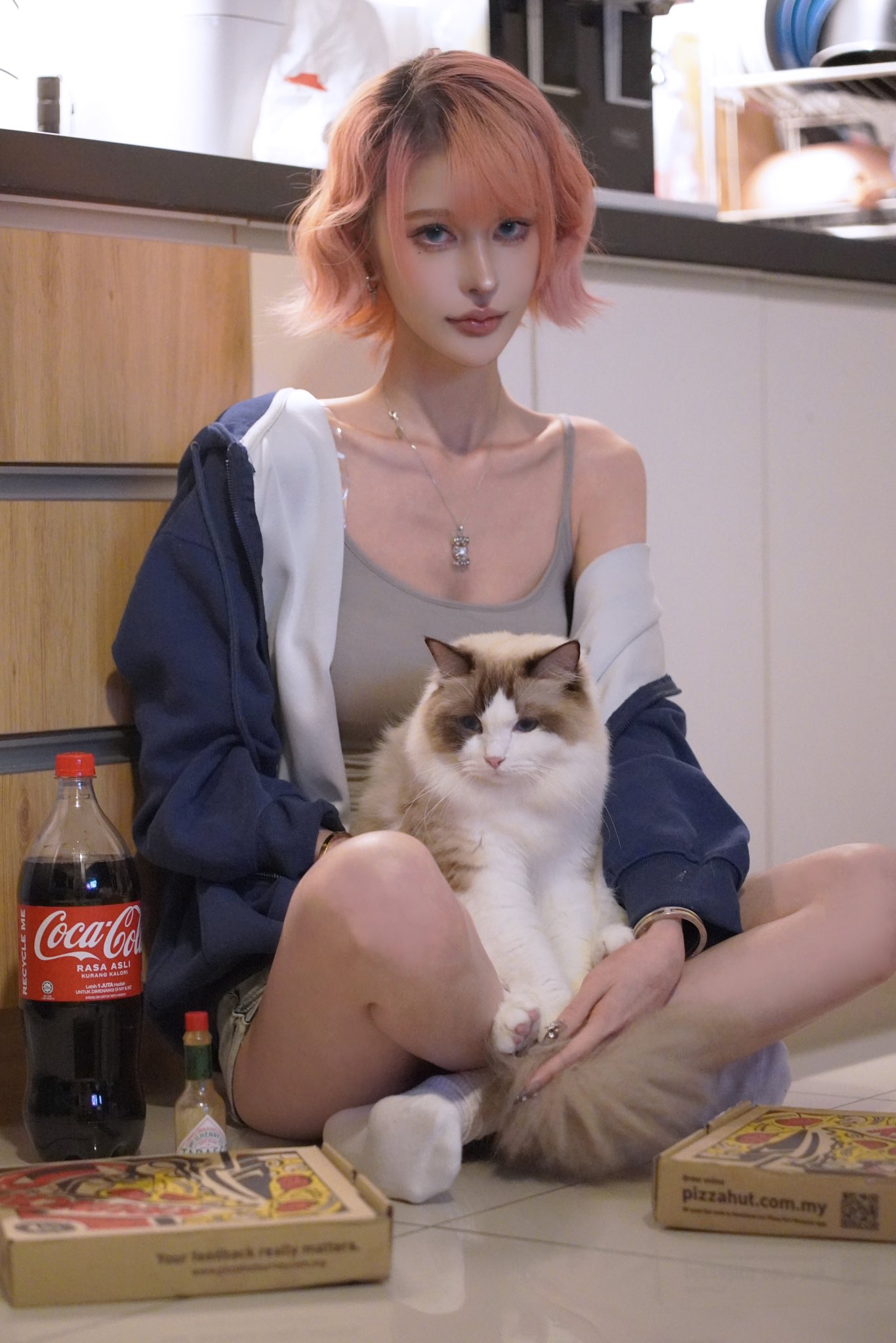 a young woman with short, pink hair is sitting on the floor of a kitchen with a ragdoll cat resting on her lap. she's wearing a dark blue sweatshirt over a light gray tank top and white socks. two pizza boxes from pizza hut and a bottle of cocacola are on the floor near her.  the overall mood is relaxed and casual.