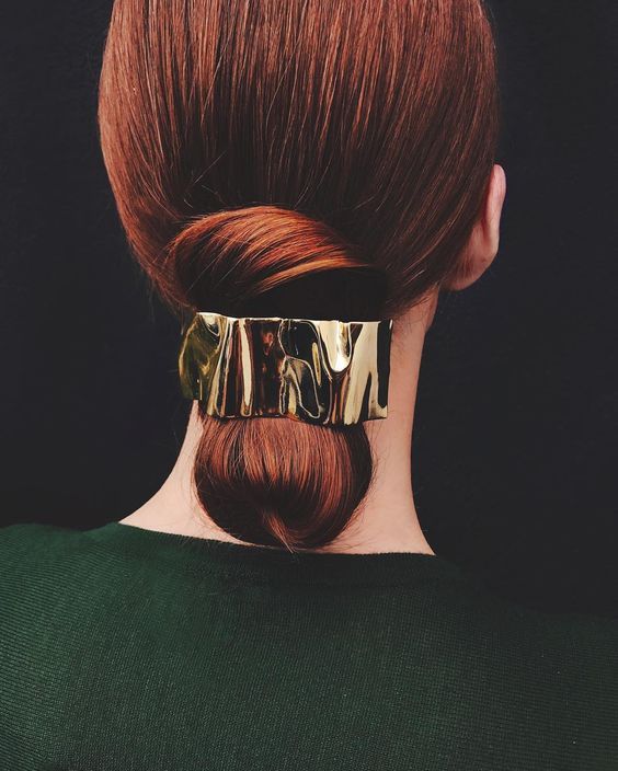 the image shows the back of a woman's head. she has long, polished, reddishbrown hair styled in a low, loose bun.  a gold, textured, rectangular hair clip is securing the bun at the nape of her neck.  she is wearing a dark emerald green sweater. the background is black, which contrasts with both her hair and the gold clip. the overall style is minimalist and elegant.