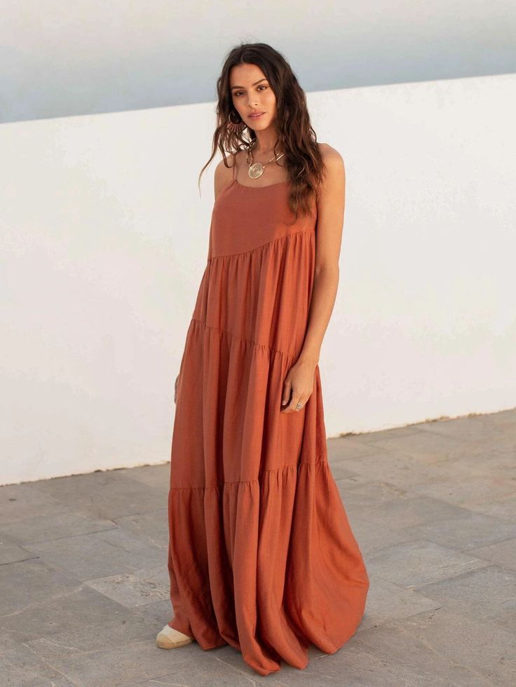 a woman stands in front of a white wall and textured stone floor, wearing a terracottacolored maxi dress with thin straps and tiered design. she has long brown hair and wears a necklace and earrings. she is also wearing espadrillestyle shoes.