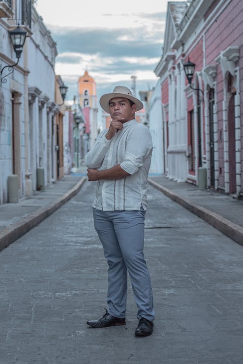 a fullbody shot of a young hispanic man in a white linen shirt and light blue pants standing in a narrow alleyway between colonialstyle buildings. he's wearing a lightcolored straw hat and has his hand on his chin in a thoughtful pose. the image is welllit, seemingly taken on a cloudy day, and the architecture of the buildings in the background adds a context to the photo. the overall style is casual and stylish.