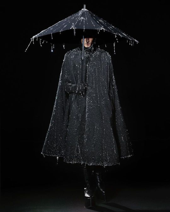 a man stands in a dark setting, wearing a black, shiny cape and holding a black umbrella with dangling accents. he is also wearing shiny black boots with platform soles.