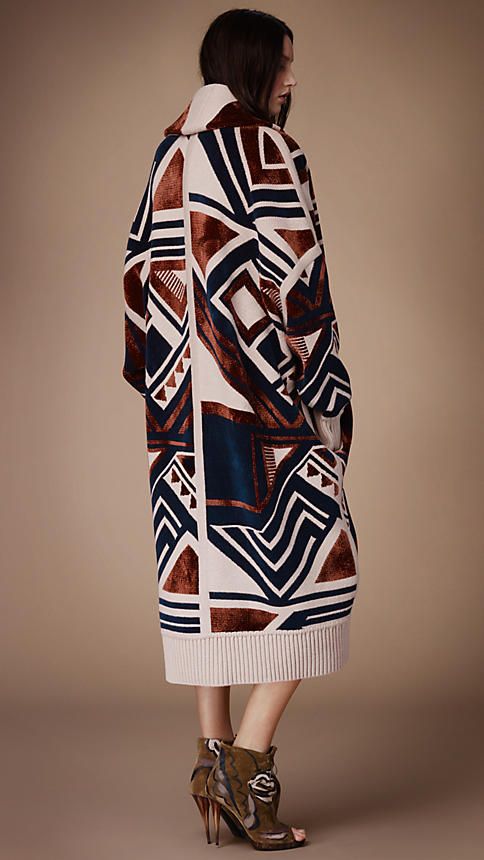 a back view of a woman's model wearing a long, geometric patterned coat. the coat is predominantly creamcolored with geometric shapes in burgundy, teal, and navy blue. the model is wearing ankle boots and is standing on a neutralcolored background.