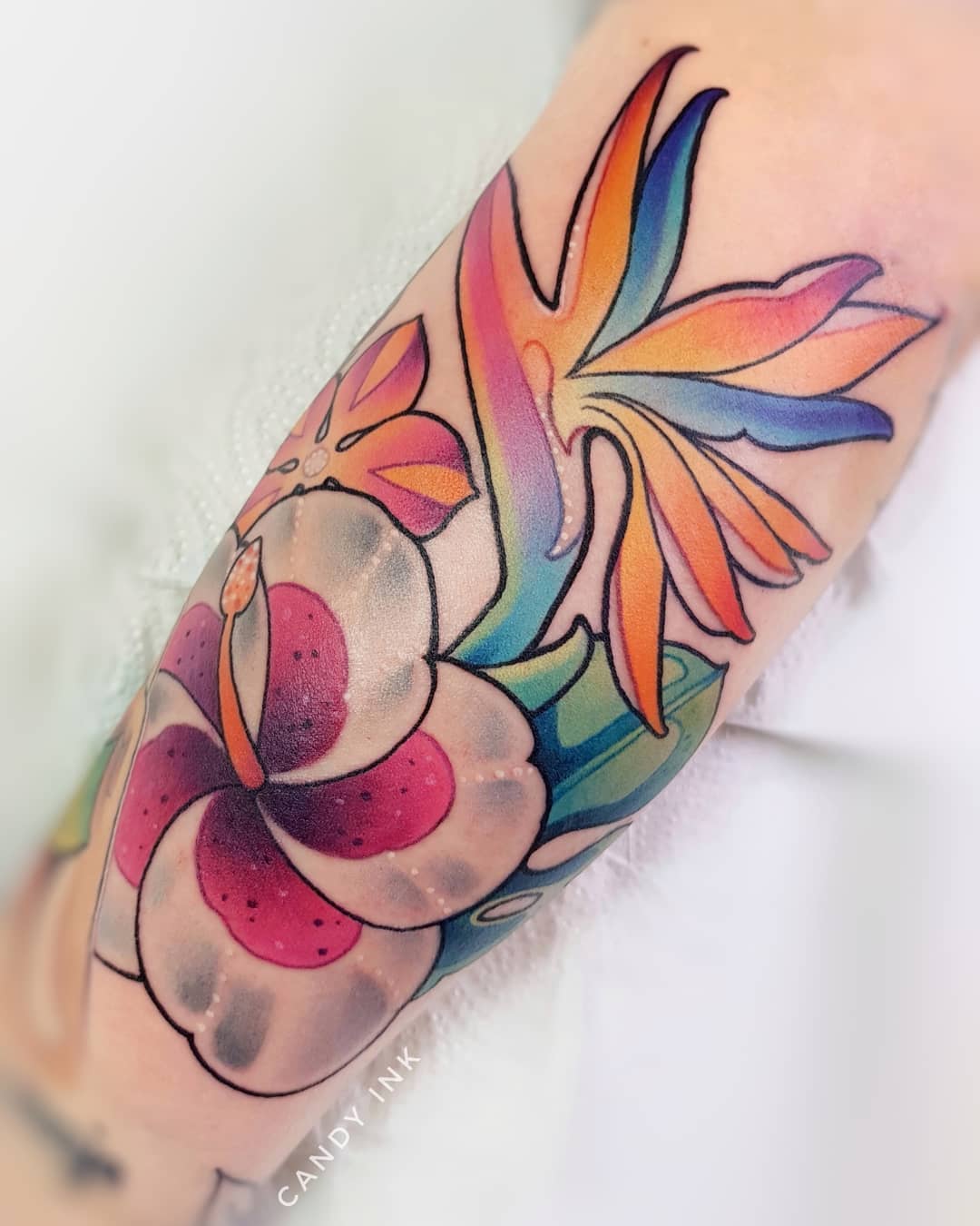 a colorful tattoo of a bird of paradise and a hibiscus flower on someone's inner arm. the tattoo is done in a vibrant, illustrative style with bold lines and shading. the bird of paradise is rendered in shades of blue, green, orange, and yellow, while the hibiscus is primarily pink and white with green leaves. the tattoo artist's signature, "candy ink," is visible in small letters below the hibiscus.