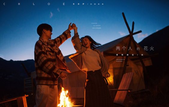 a couple is dancing in front of a bonfire at a campsite, mountains are in the background. the photo is taken at night and the main source of light comes from the bonfire. the photo is taken at eye level.