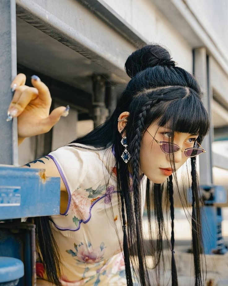 a young woman with an edgy style and long black hair styled in multiple braids is pictured outdoors in an urban, slightly industrial setting.  she is wearing a traditional style chinese top with a floral pattern in tones of cream, peach and green. she has multiple ear piercings and is wearing large earrings with blue stones. she is also wearing thin, rectangular sunglasses with a pink tint. the image is taken at eye level and uses natural light.  the overall tone of the image is slightly muted and overcast.