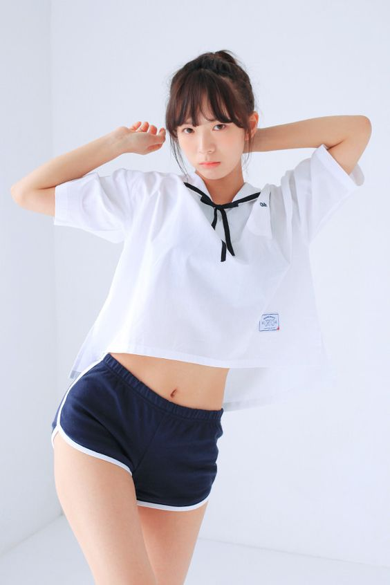 a young woman with dark hair tied back poses in a white cropped tshirt and navy blue athletic shorts. the shirt has a small logo on the front and is tied at the collar. the background is a plain white studio setting with soft, naturallooking lighting.