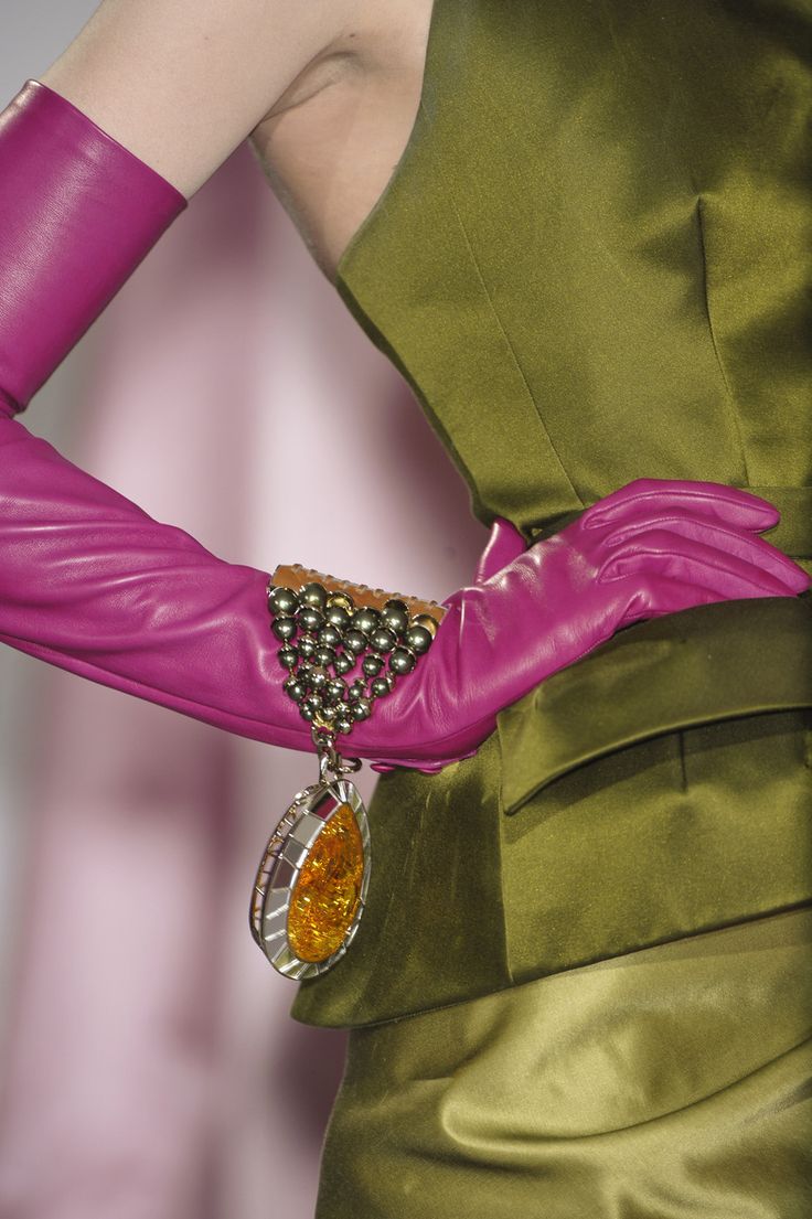 loseup shot of a woman's arm and torso, showcasing a stylish outfit.  she's wearing long, fuchsiacolored leather gloves and a flowing olive green satin dress.  a unique braceletpendant hybrid adorns her wrist, featuring a silver framework encrusted with dark beads and a teardropshaped amber stone. the image focuses on the texture of the fabrics and the detail of the jewelry, highlighting the luxurious and elegant feel of the ensemble. the background is a blurred, soft pink.