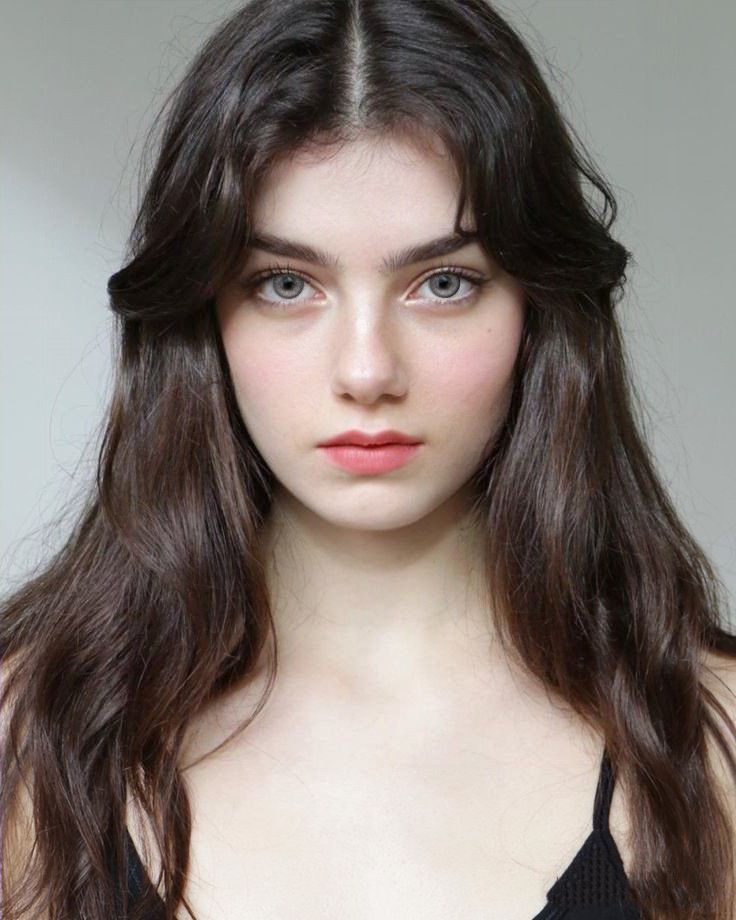the image is a closeup portrait of a young woman with long, wavy brown hair. her hair is parted in the middle and falls around her face and shoulders. she has pale skin, light gray eyes, and minimal makeup.  she's looking directly at the camera with a neutral expression. she is wearing a darkcolored spaghettistrap top. the background is plain and light gray, drawing focus to the subject.  the overall impression is one of beauty and serenity.