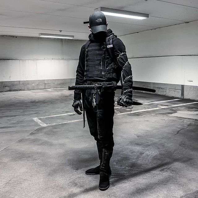 a man dressed all in black, including black clothing, a black hat, and black gloves is standing in a parking garage. he is holding a katana sword in his right hand. the image is in black and white, giving it a dark and mysterious feel.